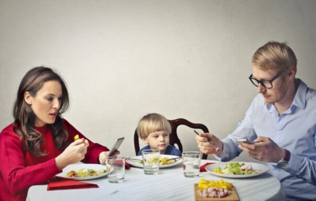 Quanto tempo de tela é demais para seus filhos? Curadoria 60 Heroes: Criança vendo seus pais verificando telefones enquanto come